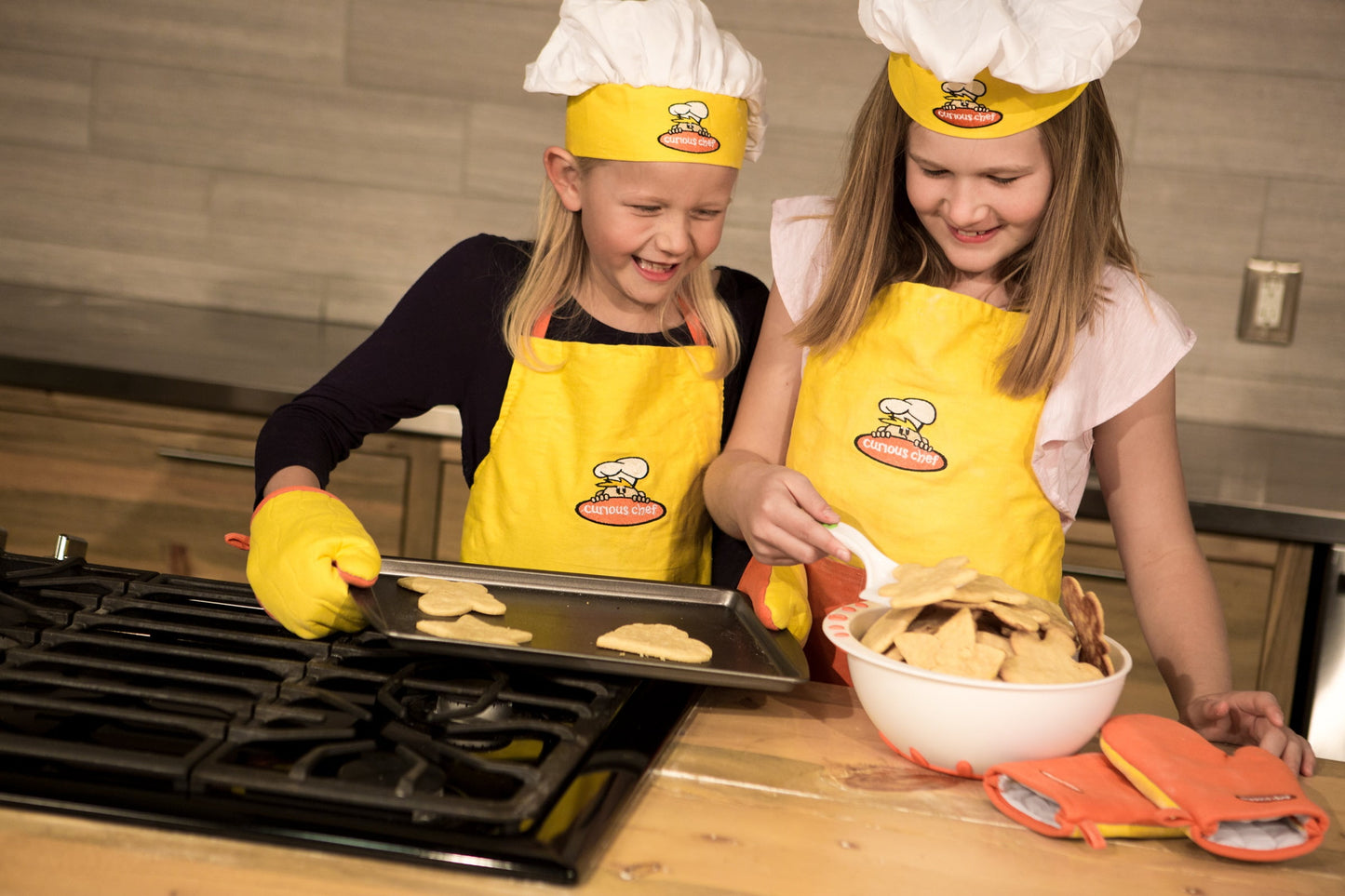 Two children modeled by girls wearing yellow aprons, white chef hats, and yellow oven mitts baking cookies on a stovetop, with a baking tray and bowl of dough