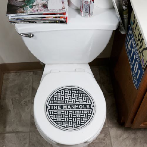 Manhole cover-style toilet seat cover with black geometric pattern and 'THE MANHOLE' text, installed on white toilet