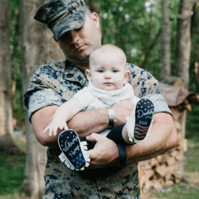 Modeled by a child wearing Baby Says Chalkboard Moccasins, held by an adult in military uniform outdoors