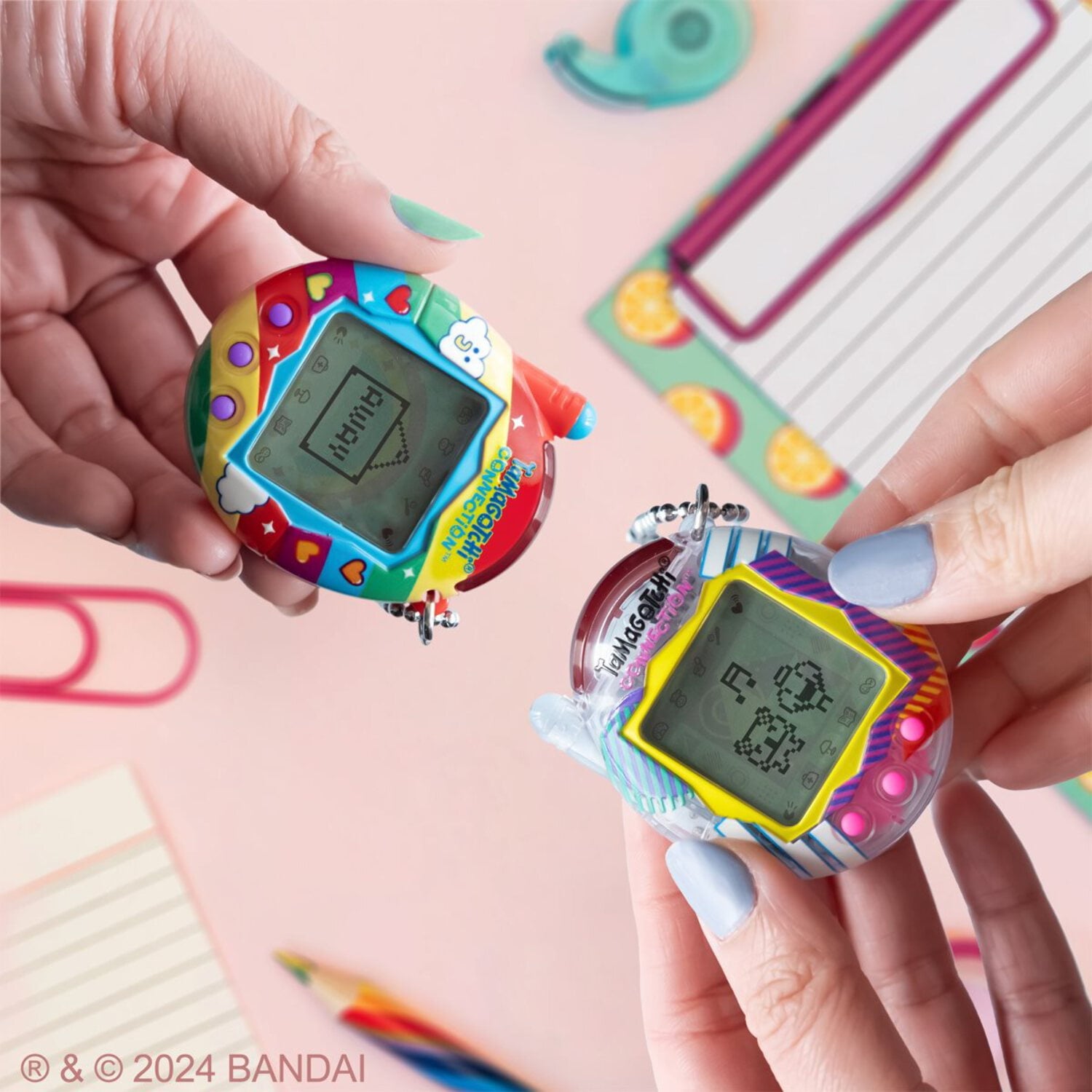 Two colorful Tamagotchi virtual pets held in hands modeled by a child, one with red and blue casing, one with yellow and purple casing, against pink background