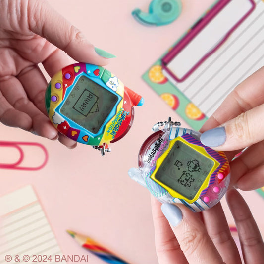 Two colorful Tamagotchi virtual pets held in hands modeled by a child, one with red and blue casing, one with yellow and purple casing, against pink background