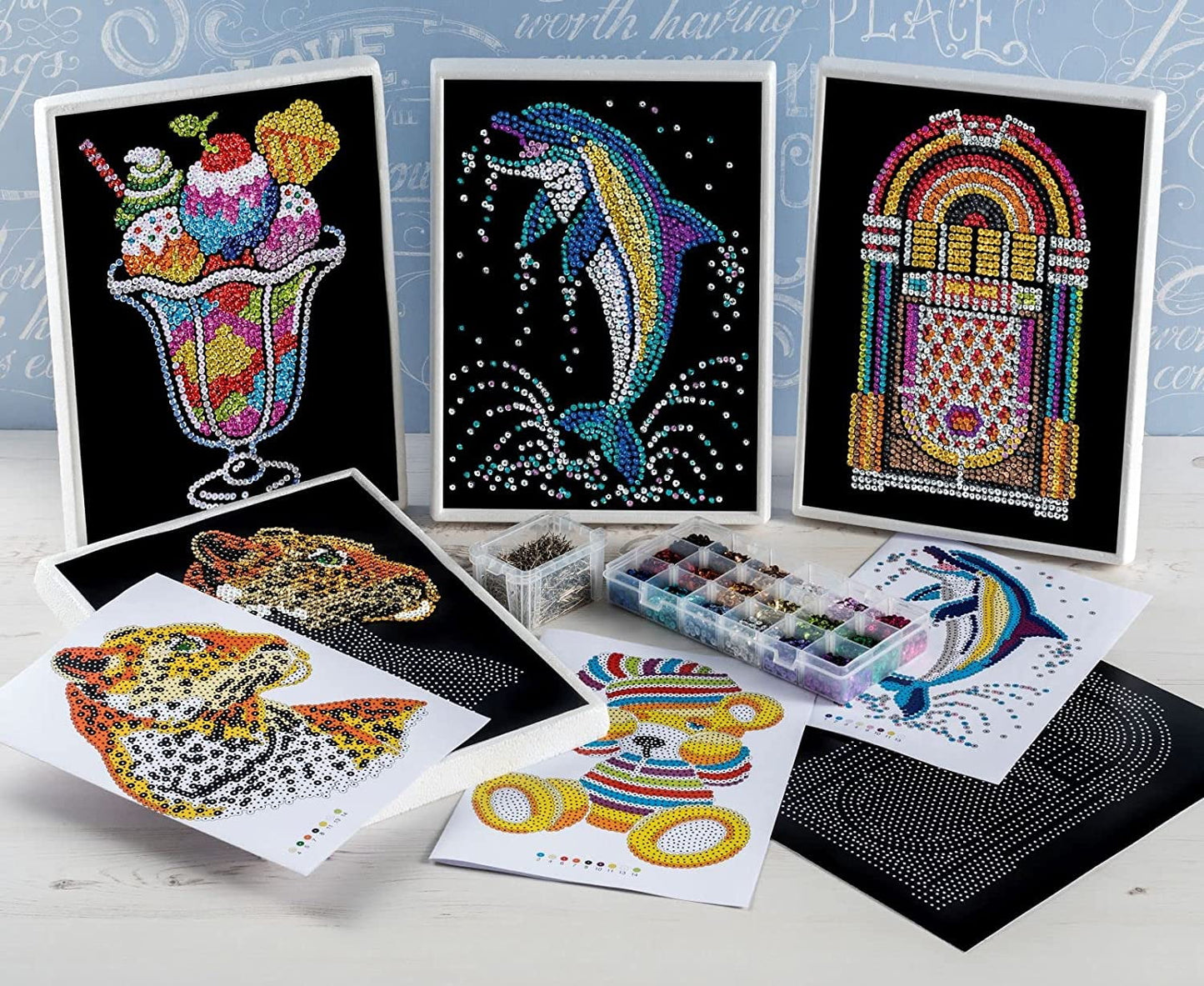 Sequin Art craft kit display showing completed colorful sequin designs including ice cream, dolphin, jukebox, fish, and llama, with instruction sheets, color-co