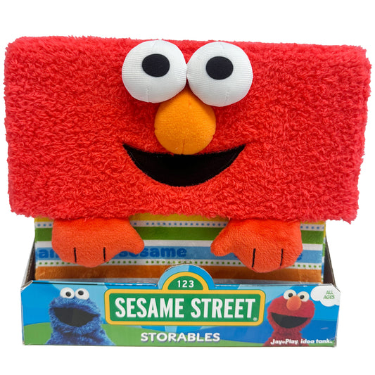 Red plush Elmo character storage bin with white eyes, orange nose, black smile, and orange arms, displayed in packaging labeled Sesame Street Storables