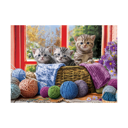 Three tabby kittens looking out a red window beside a wicker basket with colorful yarn balls and knitted blankets, purple flowers visible on right side