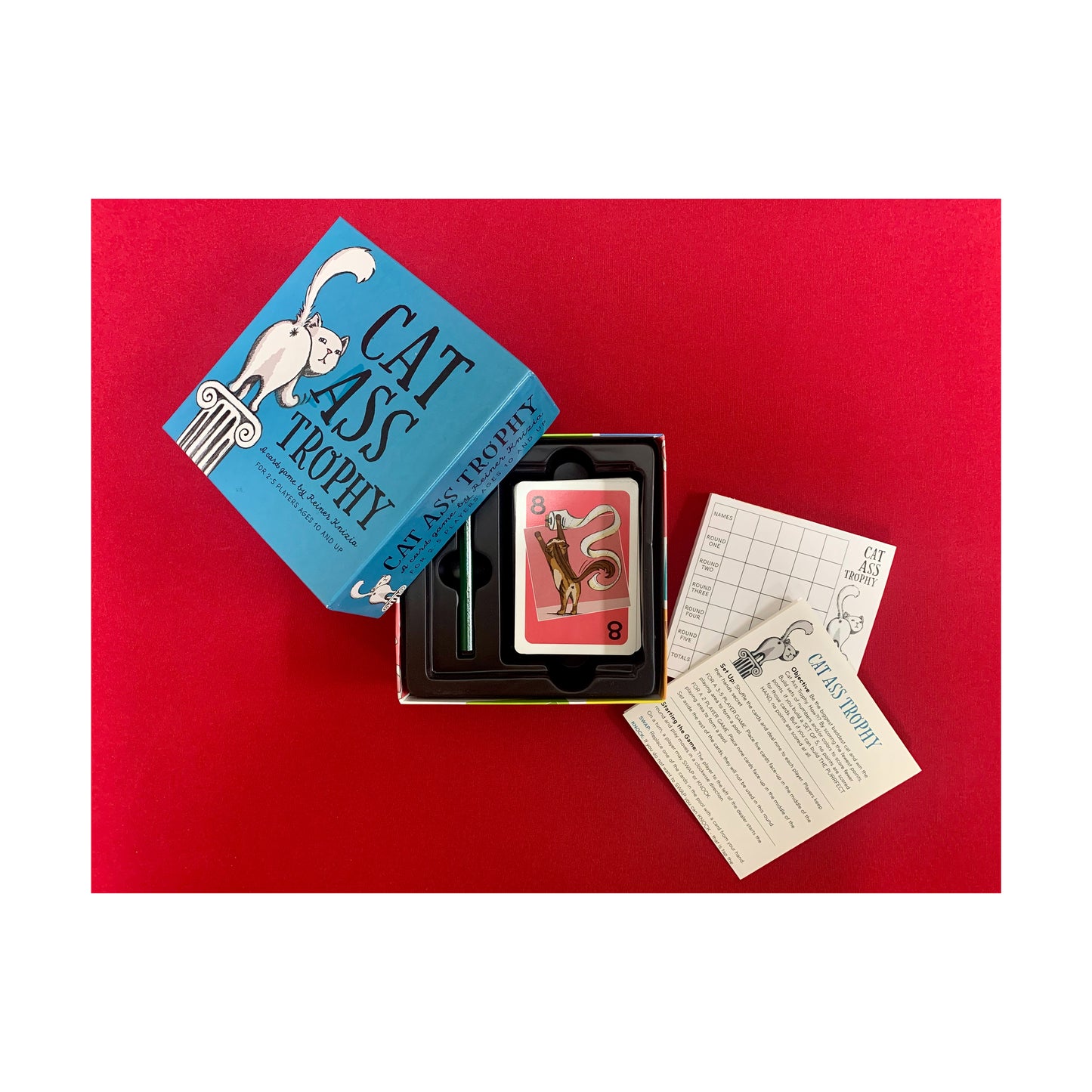 Cat Ass Trophy card game set with blue box, game cards, and instruction sheets on red background