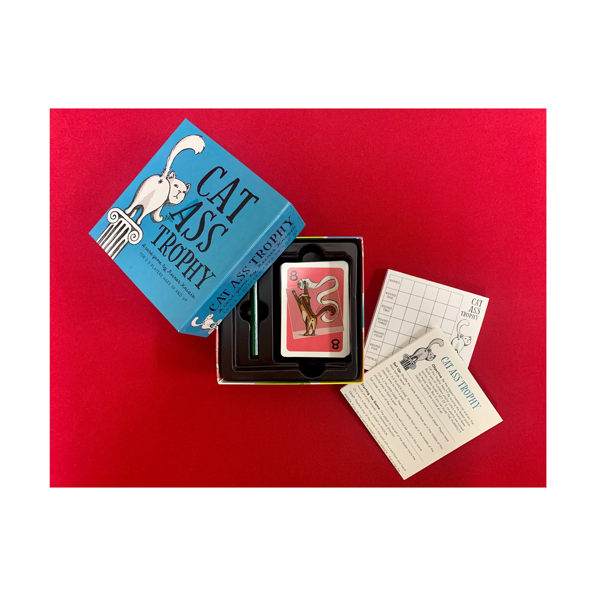 Cat Ass Trophy card game set with blue box, game cards, and instruction sheets on red background