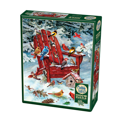 Cobble Hill Adirondack Birds 1000-piece jigsaw puzzle box featuring colorful winter birds perched on and around a red wooden chair in snow